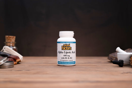 A white bottle of Natural Factors Alpha-Lipoic Acid 600 mg capsules stands on a wooden surface against a dark background, styled with small herbal props on both sides.