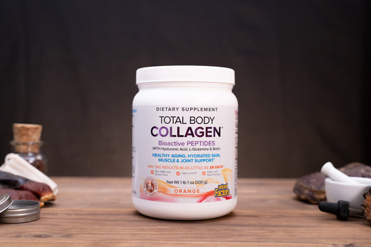 A white plastic bottle of Total Body Collagen dietary supplement with an orange flavor label, standing on a wooden surface with blurred natural props in the background.