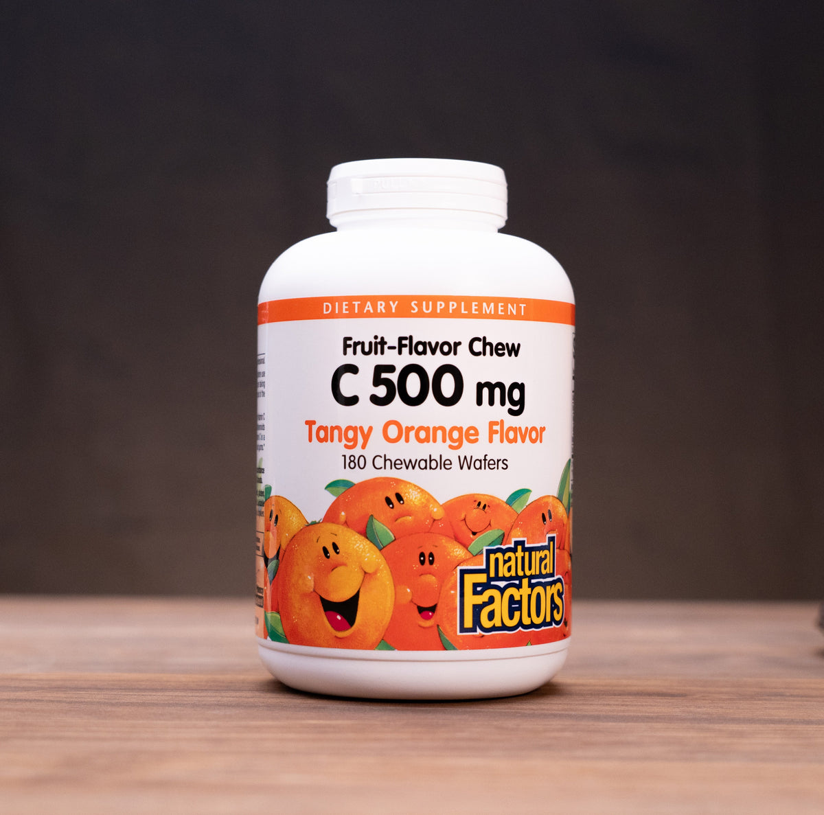 A white bottle of Natural Factors Fruit-Flavor Chew C 500 mg Tangy Orange Flavor dietary supplements, set on a wooden table with blurred herbal props in the background.