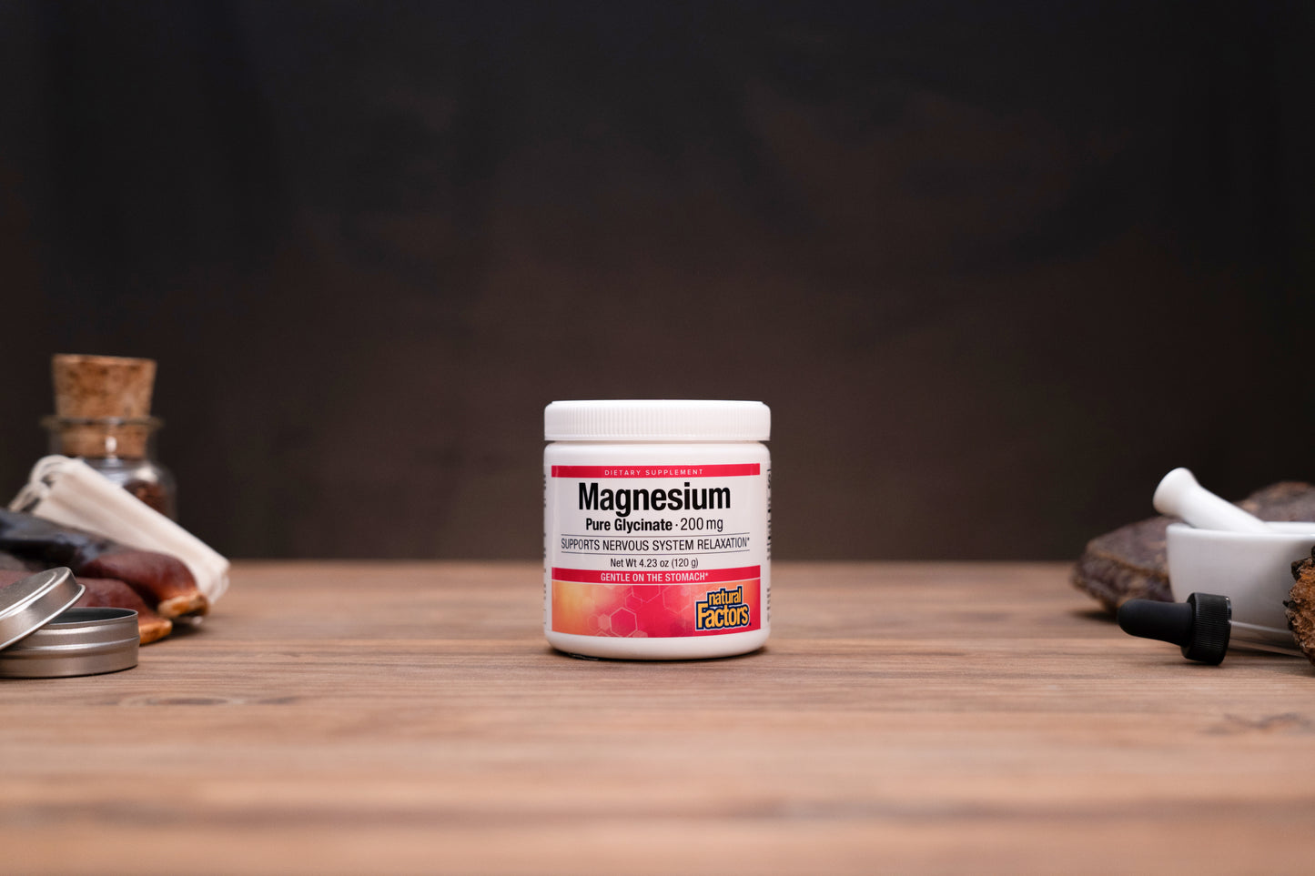 A white plastic jar of Magnesium Free Spouse supplement with a white lid, standing on a wooden surface against a dark background, flanked by blurred herbal props.