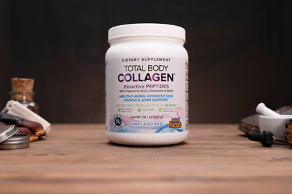 A white plastic bottle of Total Body Collagen dietary supplement, unflavored, standing on a wooden surface with a dark, moody background and blurred herbal props on either side.