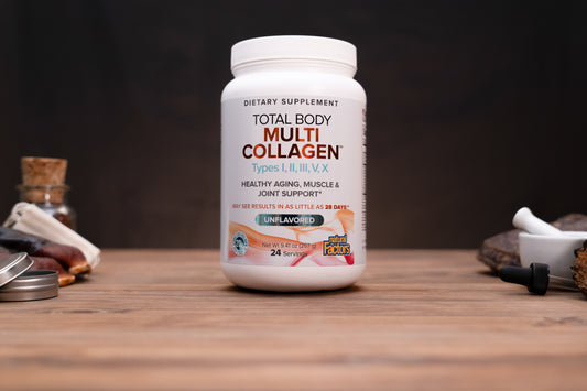A white bottle of Total Body Multi Collagen dietary supplement standing on a wooden surface against a dark background, with small blurred props on both sides.