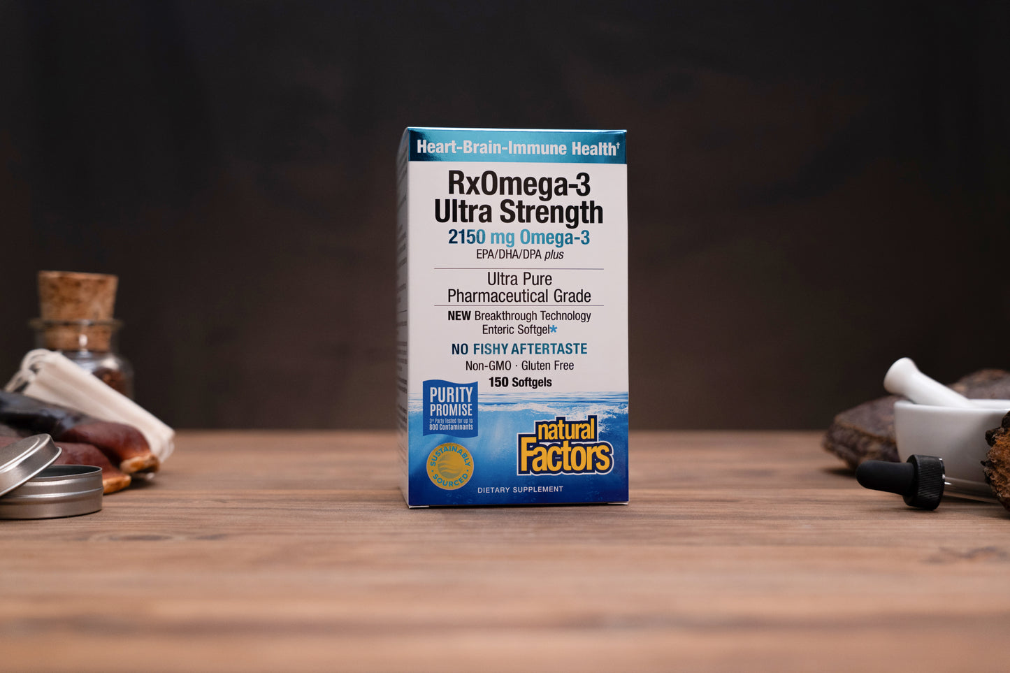 A box of Natural Factors RxOmega-3 Ultra Strength softgels stands on a wooden tabletop against a dark blurred background, with small kitchen or bar props visible on both sides.