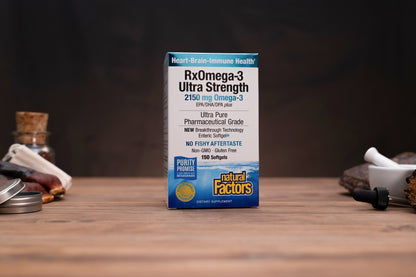 A box of Natural Factors RxOmega-3 Ultra Strength softgels stands on a wooden tabletop against a dark blurred background, with small kitchen or bar props visible on both sides.