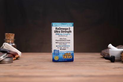 A box of Natural Factors RxOmega-3 Ultra Strength fish oil stands centered on a wooden tabletop with a dark blurred background. Small props are visible on both sides of the table.