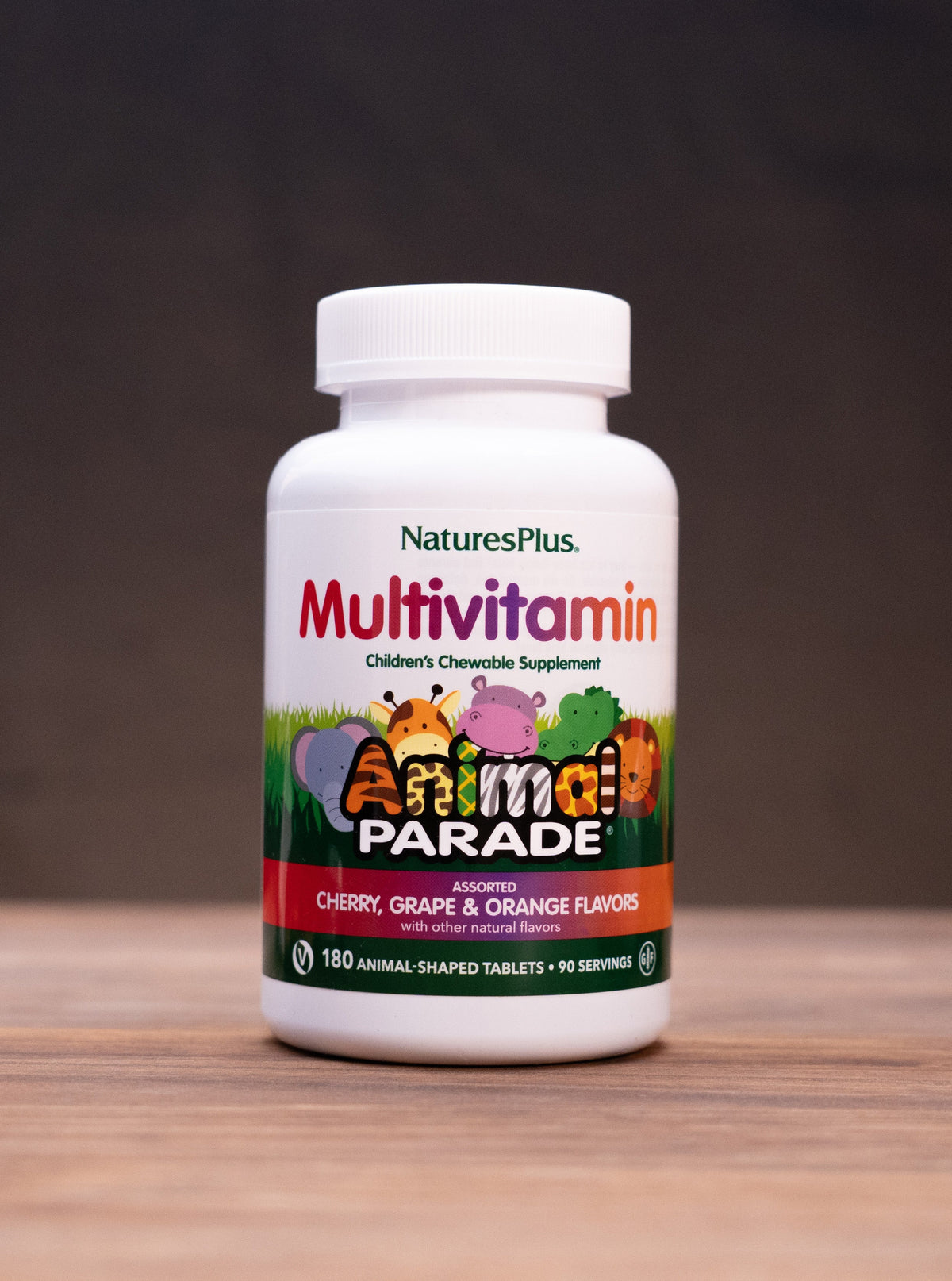A bottle of Nature's Plus Multivitamin capsules, labeled 'Animal Parade' in multicolor graphics, standing on a wooden surface with rustic props like a mortar and pestle and dried herbs blurred in the background.