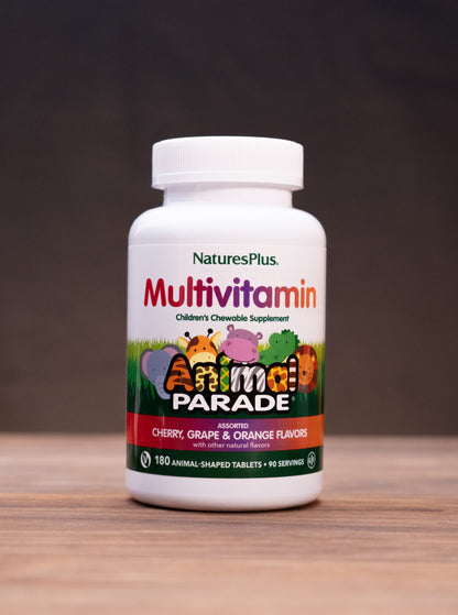 A bottle of Nature's Plus Multivitamin capsules, labeled 'Animal Parade' in multicolor graphics, standing on a wooden surface with rustic props like a mortar and pestle and dried herbs blurred in the background.