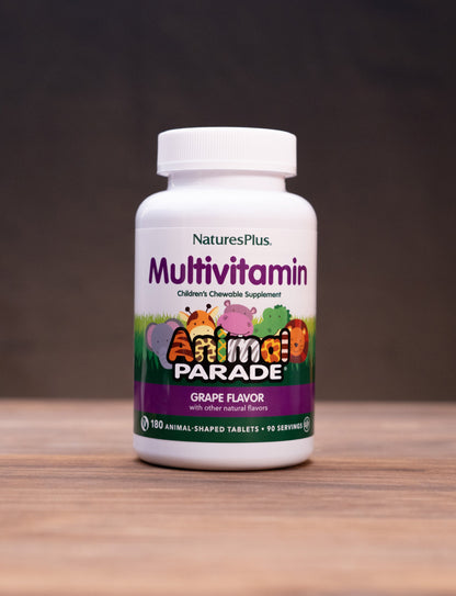 A bottle of Nature's Plus Animal Parade Multivitamin in grape flavor, standing on a wooden surface with rustic props like a mortar and pestle and dried herbs in the background.