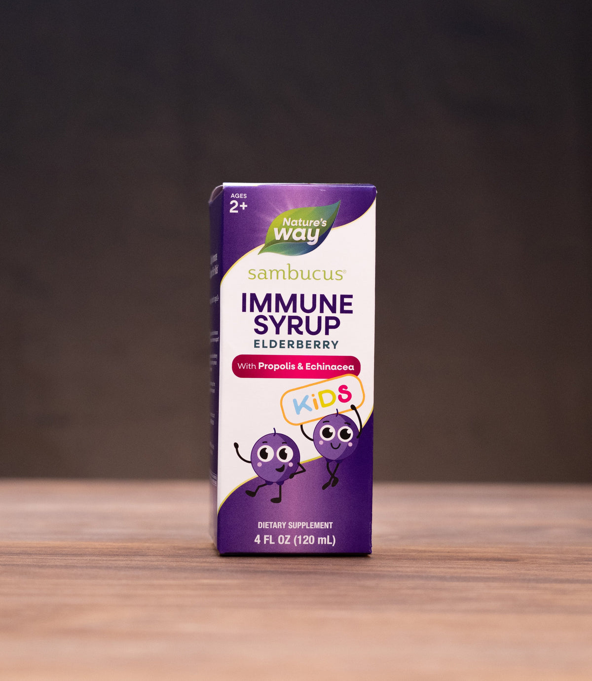 A box of Sambucus Immune Syrup Elderberry by Way Home stands on a wooden table, surrounded by blurred natural props like a cork stopper, dried herbs, and a mortar and pestle against a dark background.