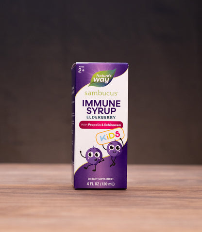 A box of Sambucus Immune Syrup Elderberry by Way Home stands on a wooden table, surrounded by blurred natural props like a cork stopper, dried herbs, and a mortar and pestle against a dark background.