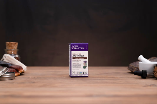 A small product box for New Chapter Herbs & Spices Compacts stands centered on a wooden tabletop against a dark blurred background, with small props visible on both sides.