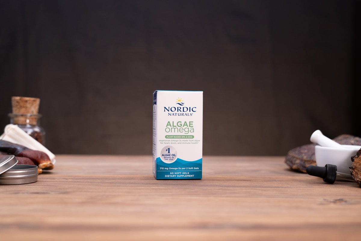 Nordic Naturals Algae Omega supplement box standing on a wooden tabletop with blurred natural props on both sides against a dark background.