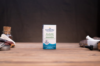 Nordic Naturals Algae Omega supplement box standing on a wooden tabletop with blurred natural props on both sides against a dark background.
