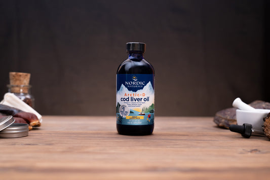 A bottle of Nordic Naturals Arctic-D cod liver oil stands centered on a rustic wooden table, surrounded by blurred natural ingredients and containers in the background.