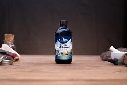 A bottle of Nordic Naturals Arctic Cod Liver Oil standing on a wooden tabletop, with blurred natural ingredients and containers in the background.