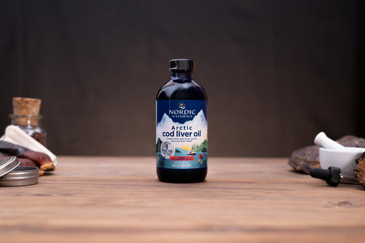 A bottle of Nordic Naturals Arctic Cod Liver Oil standing on a wooden tabletop, with blurred natural ingredients and containers in the background.