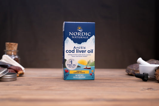 Nordic Naturals Arctic cod liver oil box on a wooden tabletop with blurred jars and a mortar in the background.