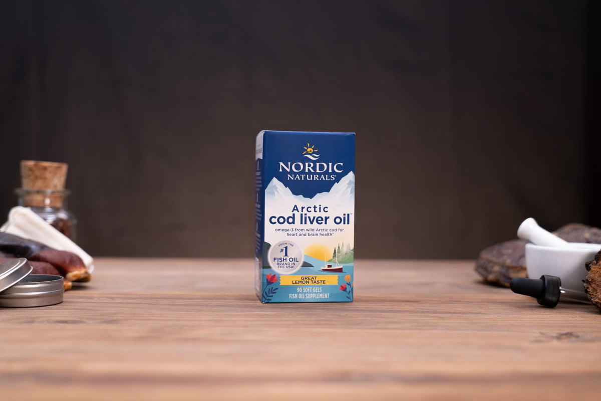 Nordic Naturals Arctic cod liver oil supplement box standing on a wooden tabletop with a dark blurred background and small props on both sides.
