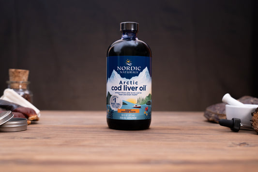A bottle of Nordic Naturals Arctic Cod Liver Oil standing on a wooden tabletop, with small kitchen or preparation props blurred in the background and foreground.