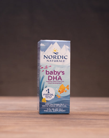 Nordic Naturals Baby's DHA 120 mg box centered on a wooden tabletop with blurred natural props on both sides against a dark background.