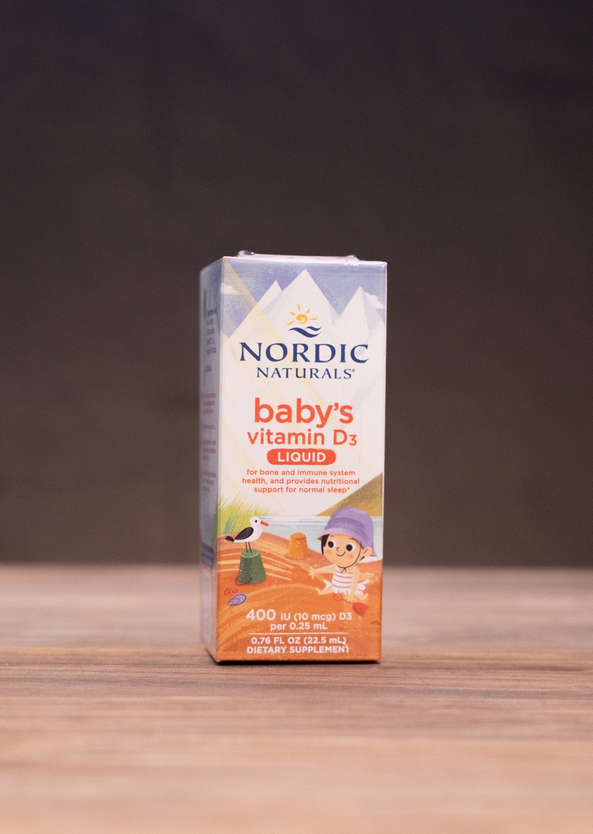 Nordic Naturals Baby's Vitamin D3 oil in a white and orange box, standing on a wooden tabletop with rustic props blurred in the background.