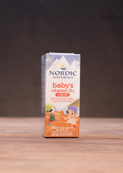 Nordic Naturals Baby's Vitamin D3 oil in a white and orange box, standing on a wooden tabletop with rustic props blurred in the background.