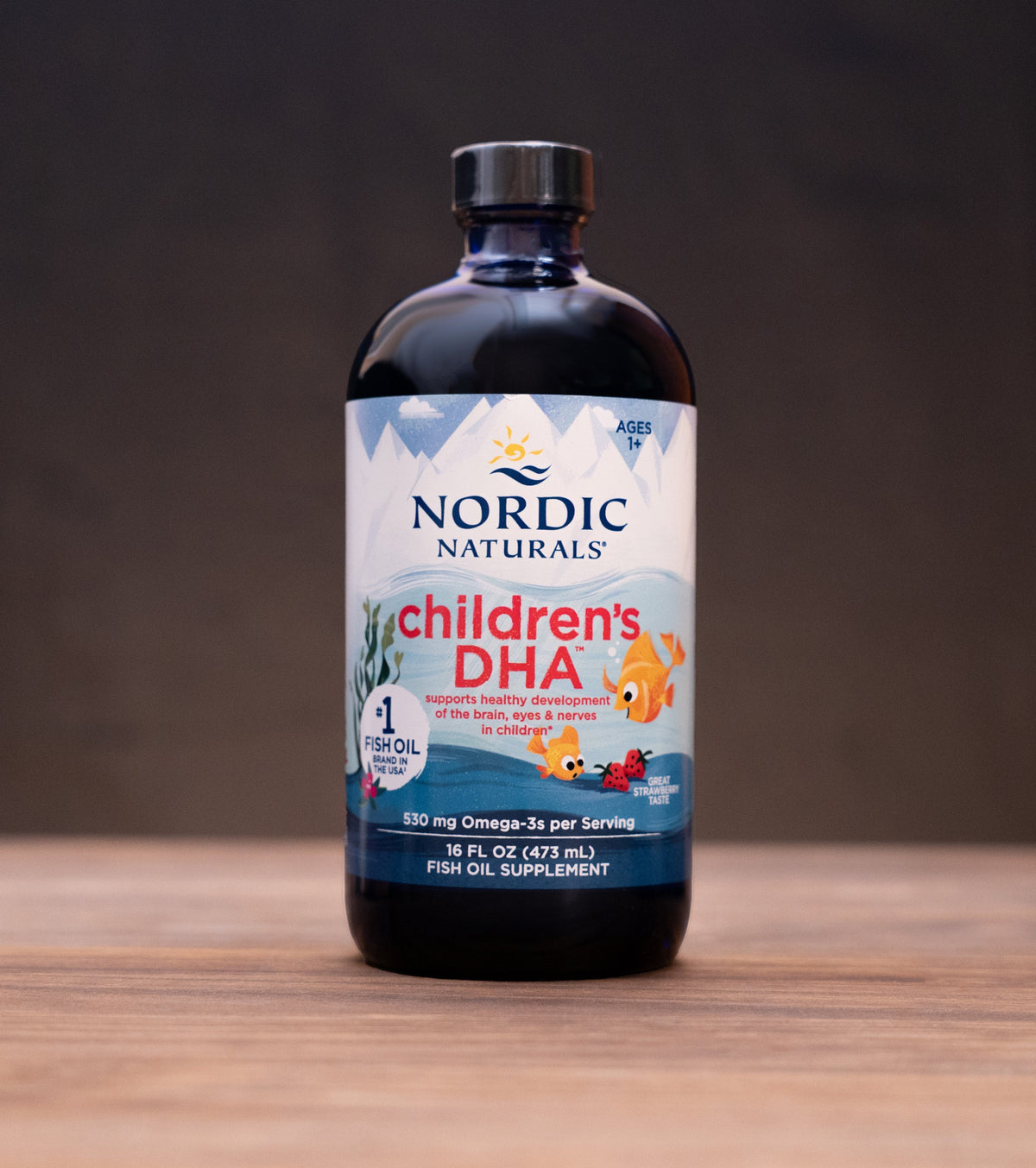 A bottle of Nordic Naturals Children's DHA dietary supplement standing on a wooden surface against a dark background, with small props visible on both sides.