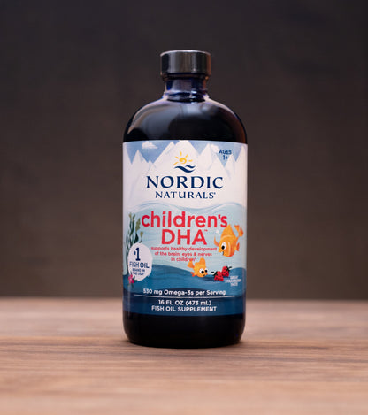 A bottle of Nordic Naturals Children's DHA dietary supplement standing on a wooden surface against a dark background, with small props visible on both sides.