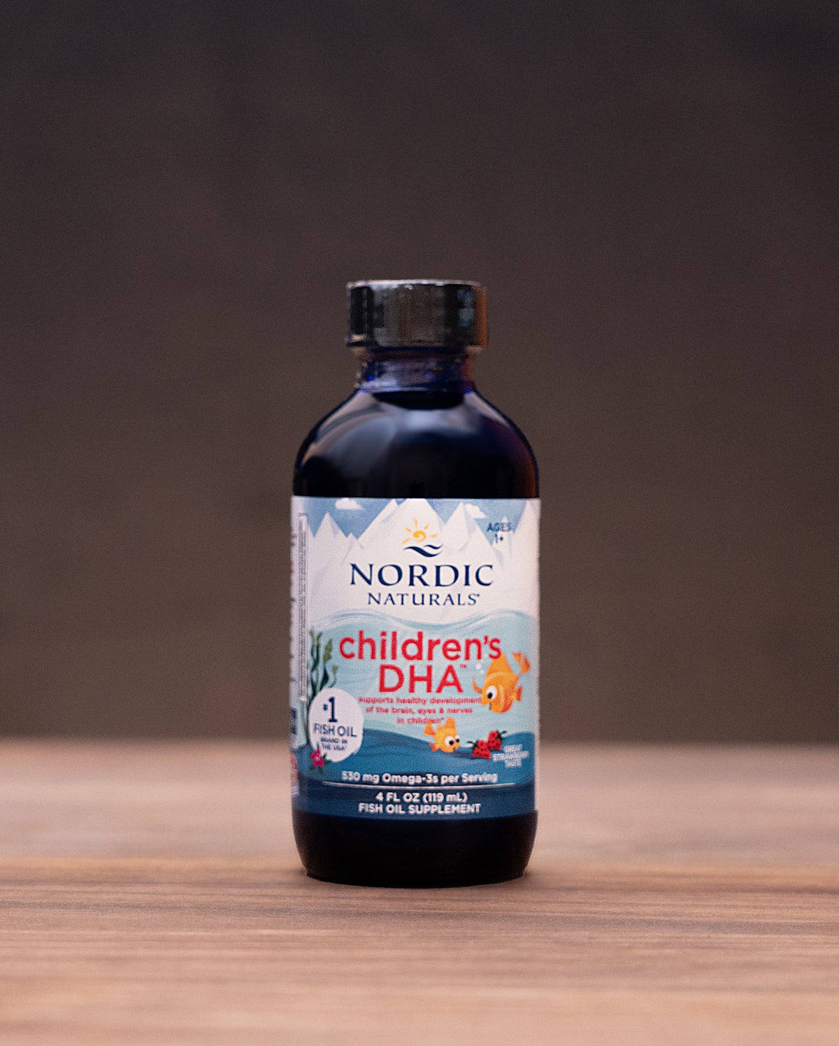 Nordic Naturals Children's DHA supplement in a dark amber glass bottle with a black cap, set on a wooden table with herbal props in the background.
