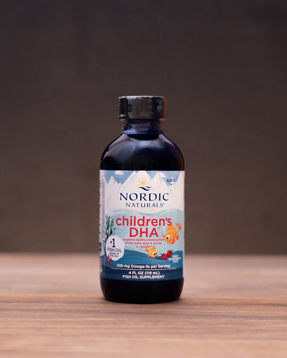 Nordic Naturals Children's DHA supplement in a dark amber glass bottle with a black cap, set on a wooden table with herbal props in the background.