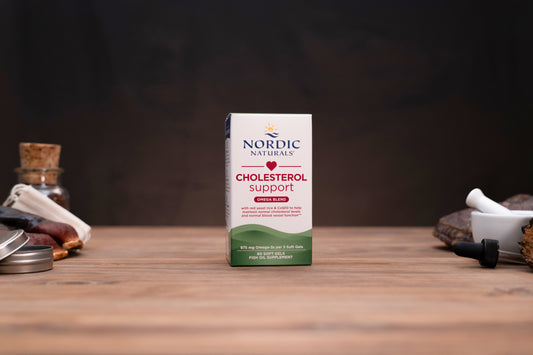 Nordic Naturals Cholesterol Support supplement box standing on a wooden tabletop with small kitchen or herbal-prep props blurred in the background.