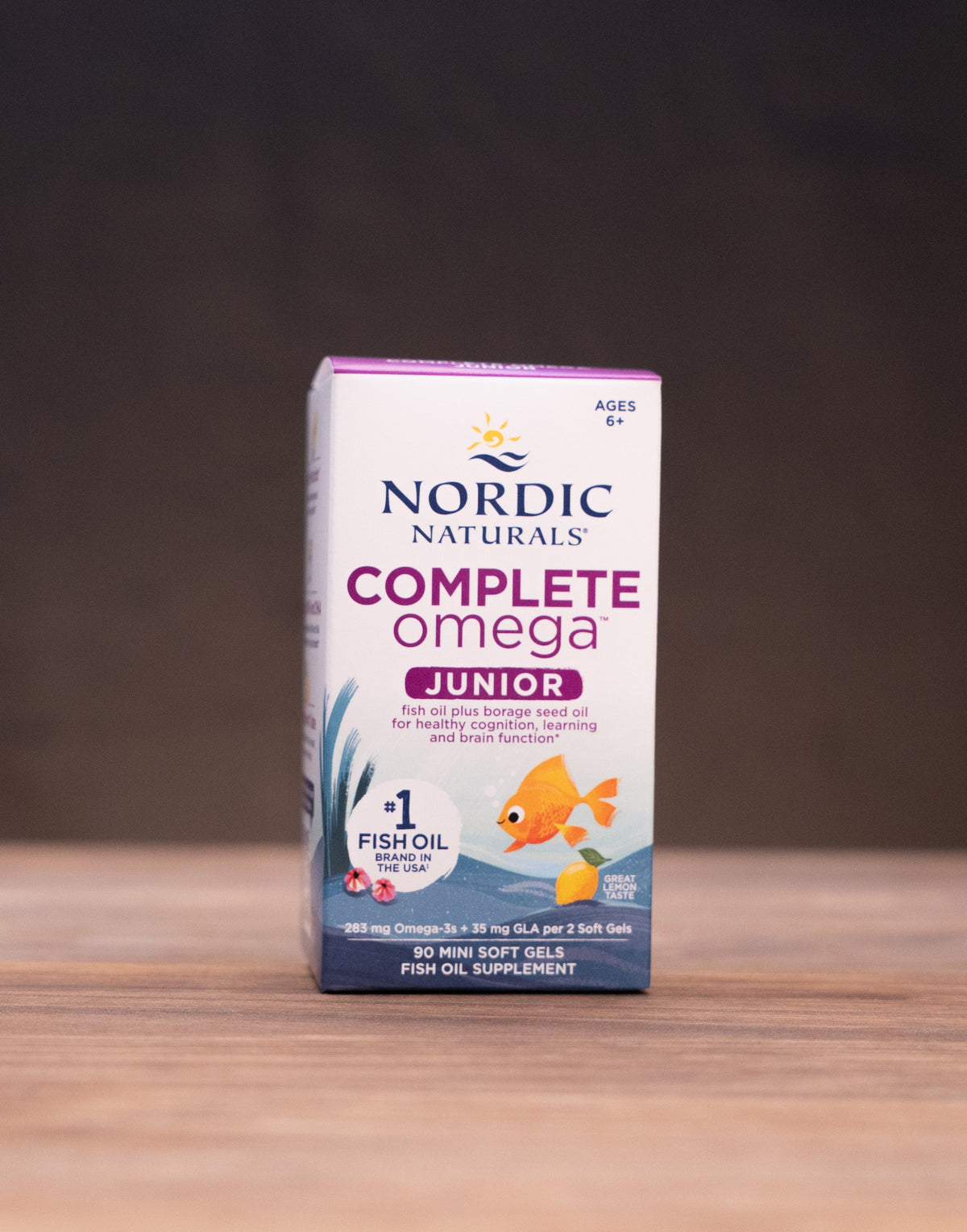 A box of Nordic Naturals Complete Omega Junior fish oil capsules stands on a wooden tabletop against a dark background, with small blurred props on both sides.