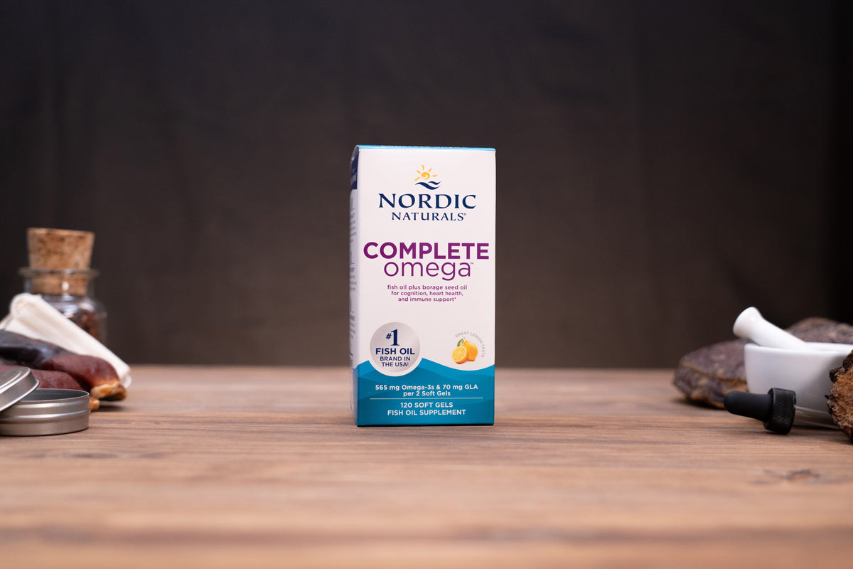 Nordic Naturals Complete Omega fish oil supplement box standing on a wooden tabletop with a dark blurred background and small props on both sides.