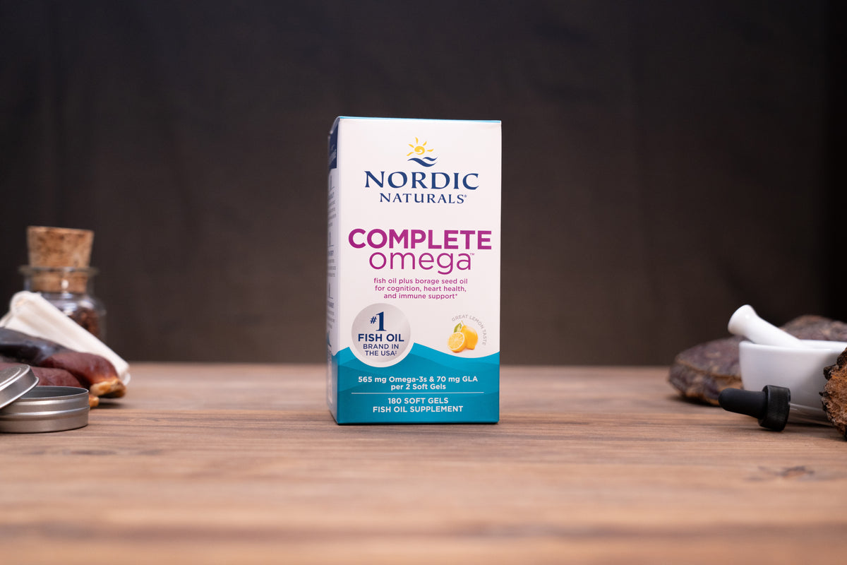 Nordic Naturals Complete Omega 3 soft gels in a white and teal box, displayed on a wooden surface with herbal props in the background.