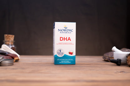 Nordic Naturals Complete Omega fish oil supplement box standing on a wooden tabletop with a dark blurred background and small props on both sides.