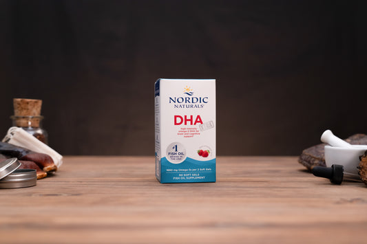 Nordic Naturals DHA fish oil supplement box standing upright on a wooden tabletop, with blurred jars and a mortar pestle in the background.