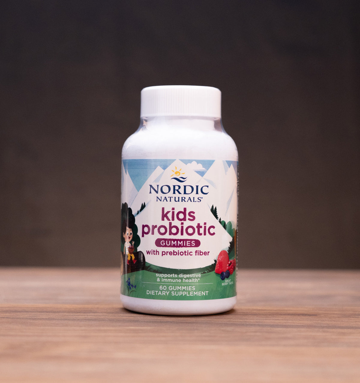 A bottle of Nordic Naturals Kids Probiotic with prebiotic fiber, set on a wooden tabletop with blurred natural props in the background.