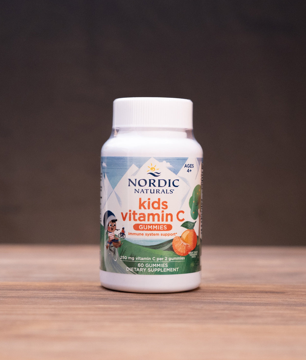 A bottle of Nordic Naturals Vitamin C dietary supplement standing on a wooden tabletop against a dark, blurred background.
