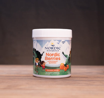 A white plastic jar of Nordic Naturals Nordic Berries (Raspberry Lemonade flavor) sits on a rustic wooden table. The background is dark and moody, with blurred natural elements like a cork stopper, dried berries, and a mortar and pestle on either side, creating a natural, outdoor-inspired aesthetic.