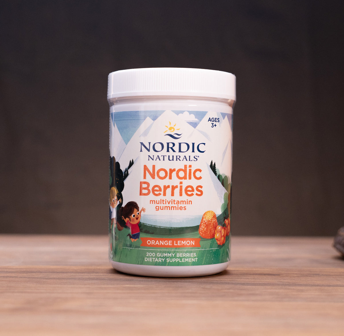 A white plastic bottle of Nordic Naturals Nordic Berries multivitamin gummies, orange lemon flavor, standing on a wooden tabletop with rustic props blurred in the background.