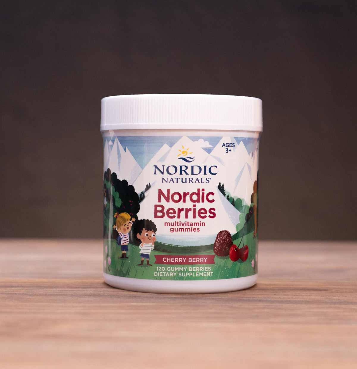 A white plastic jar of Nordic Naturals Nordic Berries (Cherry Berry flavor) centered on a wooden table, with rustic herbal props blurred in the background.