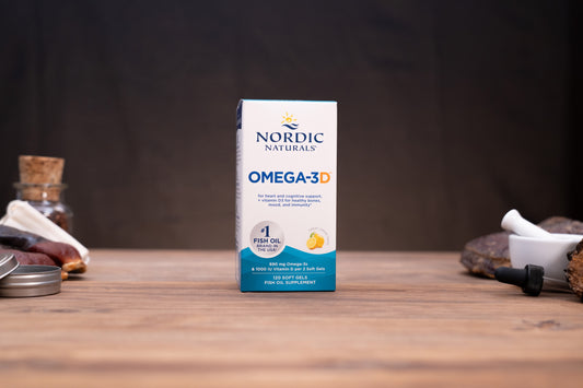 Nordic Naturals Omega-3 3D fish oil supplement box standing on a wooden tabletop with blurred jars and a white mortar in the background.