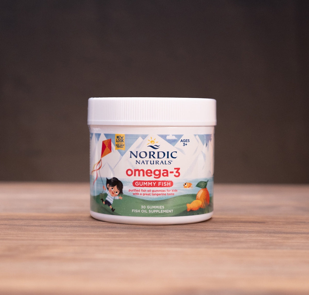A white jar of Nordic Naturals Omega-3 fish oil supplement, displayed on a wooden surface with a dark, moody background and subtle herbal props.