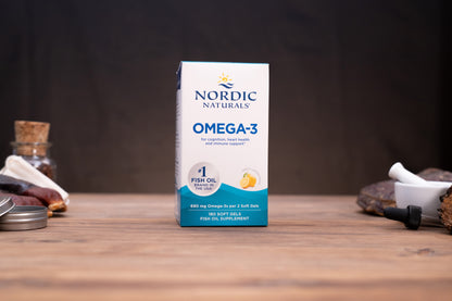 Nordic Naturals Omega-3 fish oil supplement box standing on a wooden tabletop with a dark blurred background and small props on both sides.