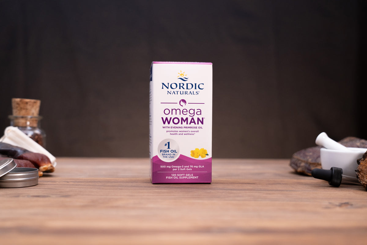 Nordic Naturals Omega Woman dietary supplement box standing on a wooden tabletop with blurred jars and a mortar in the background.