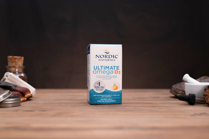 Nordic Naturals Ultimate Omega D3 fish oil supplement box standing on a wooden tabletop against a dark background, with small props visible on both sides.