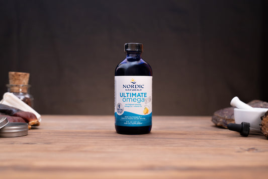 A bottle of Nordic Naturals Ultimate Omega dietary supplement centered on a wooden tabletop, with blurred natural props on both sides against a dark backdrop.