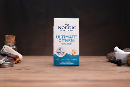 Nordic Naturals Ultimate Omega fish oil supplement box standing on a wooden surface with blurred jars and a mortar and pestle in the background.