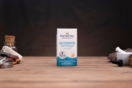 Nordic Naturals Ultimate Omega dietary supplement box standing on a wooden tabletop, with small jars and a mortar-and-pestle blurred in the background.