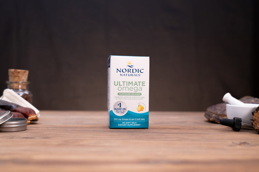Nordic Naturals Ultimate Omega dietary supplement box standing on a wooden tabletop with blurred jars and a white mortar in the background.